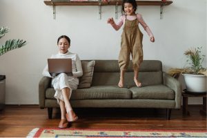 Mother sitting on the couch working on a laptop and the kid playing beside her.